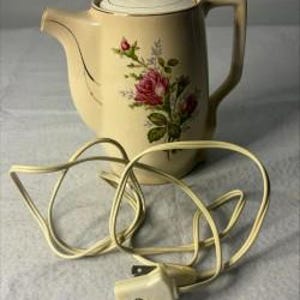 May include: A cream-colored electric ceramic teapot featuring a floral design of pink roses and green leaves on the front. The teapot has a matching lid, a curved handle, a spout, and a long white electrical cord with a two-prong plug attached.