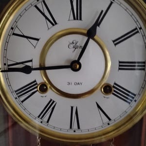 May include: A round wall clock with a gold-toned metal frame and a white face featuring black Roman numerals. The center displays the brand name Elgin in script and the text 31 DAY. Two black hands indicate the time, with two winding holes below.