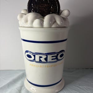 Op de afbeelding: Een witte keramische Oreo-koekjespot met een blauwe rand en het Oreo-logo. De deksel is een grote Oreo-koek, en de bovenkant van de pot is ontworpen om op slagroom te lijken. De tekst "America's Favorite Cookie" staat onder het logo.