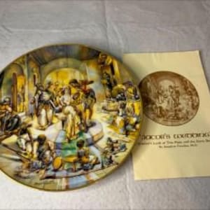 May include: A round decorative ceramic plate featuring a colorful, detailed illustration of a wedding scene with multiple figures in an architectural setting. Beside it lies a cream-colored paper pamphlet titled "Doctor's Wedding" with a sepia-toned image.