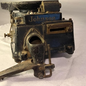May include: A vintage black metal Johnson machine featuring a prominent horizontal slot, a circular top component, and a protruding lower arm mechanism. The word Johnson is embossed in gold lettering on the front of the dark, weathered casing.