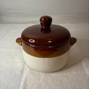 Op de afbeelding: Een ronde keramische bonenpot met een tweekleurig ontwerp, voorzien van een glanzend donkerbruin deksel en bovenrand, overgaand in een crèmekleurige basis. De pot heeft twee kleine handvatten aan de zijkanten en een knop op het deksel.
