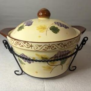 May include: A cream-colored ceramic casserole dish with a matching lid, featuring painted purple grape clusters and green leaves. The dish has brown side handles and a brown knob, resting in a black wrought iron stand with decorative scrollwork.