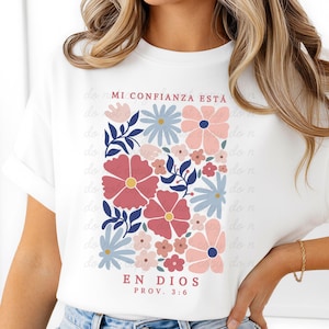以下が含まれることがあります： ピンク、青、赤の色合いの花柄が特徴の白いTシャツ。「MI CONFIANZA ESTÁ EN DIOS PROV. 3:6」の文字が入っています。クルーネックと半袖のクラシックなデザインです。