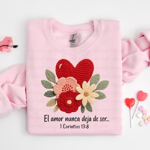 Może przedstawiać: Jasnoróżowa bluza z czerwonym szydełkowym sercem i kwiatowym wzorem. Wzór zawiera różowe, czerwone i kremowe kwiaty z zielonymi liśćmi. Tekst "El amor nunca deja de ser... 1 Corintios 13:8" jest wydrukowany poniżej.