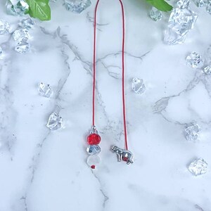 May include: A red beaded bookmark with a silver elephant charm. The bookmark features red, silver, and white beads. The elephant charm has red accents. The bookmark is on a red string.
