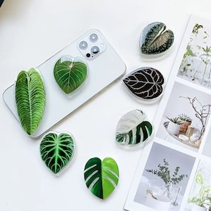 May include: Assortment of seven phone grips shaped like green leaves. The grips are made of clear plastic with a green leaf design inside. The leaves are in various shapes and sizes.