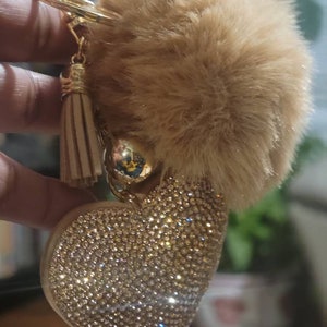 May include: A gold heart-shaped keychain with a sparkly, rhinestone design. A fluffy, brown pom-pom charm hangs from the keychain.