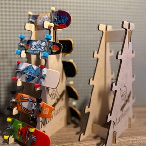 May include: A wooden display stand holding several miniature skateboards. The skateboards have various colorful designs and graphics. The stand is made of light-colored wood and has the word "Marians" engraved on it.