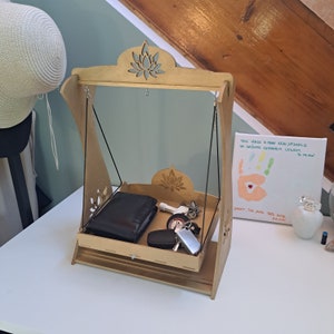 May include: A wooden swing shelf with a decorative lotus flower design. The shelf has a drawer and is holding a black wallet, keys, and a small white object. The shelf is sitting on a white table.
