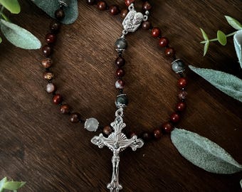 May include: A rosary with dark brown and gray beads and a silver crucifix. The beads are interspersed with gray and light-colored beads. The crucifix features a detailed depiction of Jesus Christ.