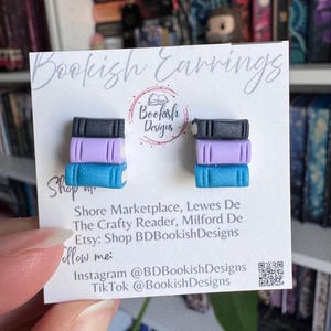 May include: A pair of stud earrings shaped like miniature stacks of books. Each earring features three stacked book shapes in black, lavender, and teal. The card reads "Bookish Earrings" and "Bookish Designs".