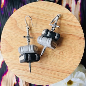 May include: A pair of silver sword earrings with stacked book charms. The books are black and gray with silver accents. The swords are detailed with intricate designs.