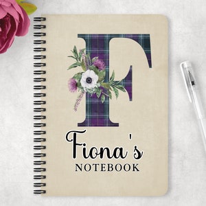 May include: A spiral bound notebook with a beige cover featuring a large purple and green plaid letter F with white flowers and green leaves. The text "Fiona's Notebook" is printed below the letter.