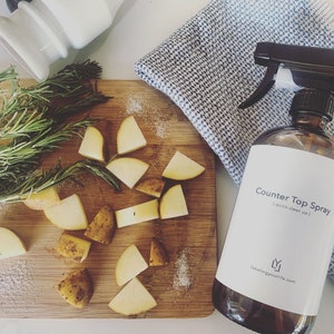 Könnte beinhalten: Eine braune Glasflasche mit einem weißen Etikett, auf dem "Counter Top Spray | quick clean up | labelingyourlife.com" steht. Die Flasche steht auf einem Holzbrett mit Kartoffeln und Rosmarin.