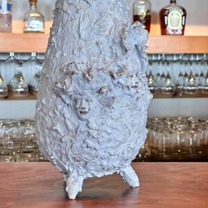 Peut inclure: Un vase en céramique blanche avec une surface texturée et un visage sculpté sur le devant. Le vase a trois pieds et est posé sur une surface en bois.