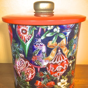 May include: A red and gold Christmas cookie tin with a festive design of ornaments, bells, and holly. The tin has a lid with a gold knob.