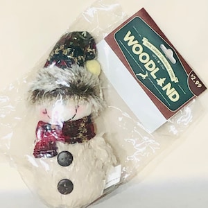Pode incluir: Um enfeite de boneco de neve de pelúcia com um chapéu xadrez, guarnição de pele marrom e um cachecol xadrez vermelho e marrom. O enfeite tem dois botões pretos e um rosto branco. O enfeite é embalado em um saco de plástico transparente com um rótulo Woodland marrom e verde que diz "Sua maior loja de Natal".