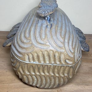 May include: A ceramic lidded container with a textured, organic design. The container is primarily blue and tan, with a lid featuring a stylized floral element. The body of the container has a repeating, curved pattern.