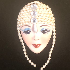 May include: A white porcelain mask with a pearl and rhinestone headpiece. The mask has blue eyes, red lips, and a white pearl nose.