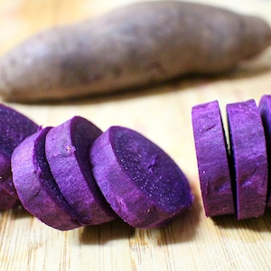 May include: Close-up of sliced purple sweet potatoes on a wooden cutting board. The vibrant purple flesh contrasts with the light brown of the board. A whole sweet potato is in the background.