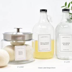 May include: Three glass bottles with labels for laundry products. The bottles are filled with clear liquids. The labels are white with black text. The text on the labels reads "Free & Gentle Detergent", "Chlorine Bleach", and "Wool Dryer Balls". The bottles are sitting on a white surface. The bottles are 3 inches by 3.75 inches and 2 inches by 2.625 inches.