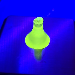 Könnte beinhalten: Ein neongrüner Flaschenverschluss mit rundem Boden und ausgestelltem Oberteil. Der Verschluss ist vor einem dunkelblauen Hintergrund beleuchtet und erzeugt einen Leuchteffekt. Das Objekt scheint aus Glas oder einem ähnlichen durchscheinenden Material zu bestehen.