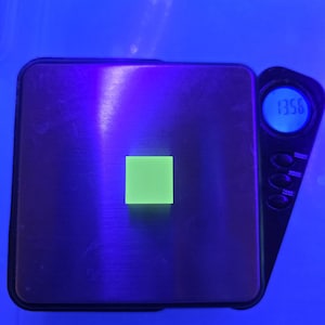 May include: A digital scale with a black base and a small, bright green square on top. The display shows a weight of 13.56. The background is a vibrant blue.