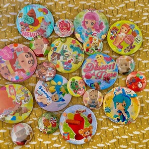 May include: A collection of colorful, round pinback buttons with various cartoon character designs. Some buttons feature text like "Dessert Time," "Angel," and "Sonny." The buttons are arranged on a yellow floral patterned surface.