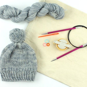 May include: A gray and white knit hat with a pom pom on top, a set of circular knitting needles, a yarn winder, and a bag of gray and white yarn.