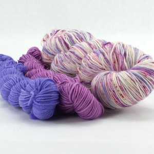 May include: A collection of yarn skeins in various shades of purple and white. The yarn is arranged in a cluster, with the colors ranging from deep violet to pale lavender and speckled white. The yarn is likely for knitting or crocheting projects.