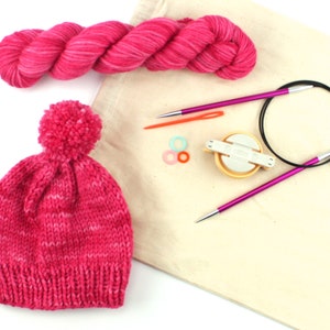 May include: A pink and white striped knit hat with a pom pom on top, a skein of pink yarn, a pair of circular knitting needles, a set of stitch markers, and a knitting needle gauge.