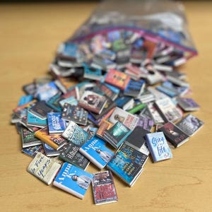 May include: A collection of miniature books, many with colorful covers and titles like "Anna" and "Story of the Life", spilling from a clear plastic bag onto a light brown surface. The books vary in size and design.