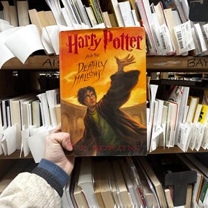 May include: A hardcover copy of "Harry Potter and the Deathly Hallows" by J.K. Rowling. The book cover features an illustration of Harry Potter with his wand raised, set against an orange and yellow background. The book is held in a library setting.