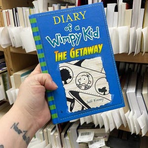 May include: A blue hardcover book titled Diary of a Wimpy Kid The Getaway by Jeff Kinney. The cover features a green and blue striped spine, yellow text, and a central illustration of a cartoon character in a circular frame on a torn paper graphic.