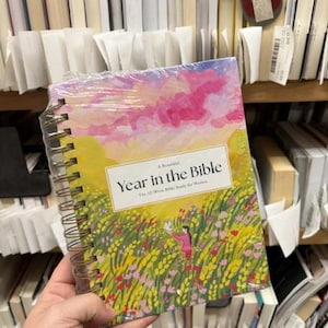May include: A spiral-bound book titled A Beautiful Year in the Bible, The 52-Week Bible Study for Women. The cover features a colorful illustration of a person in a field of yellow and pink flowers under a pink and yellow sky, wrapped in plastic.