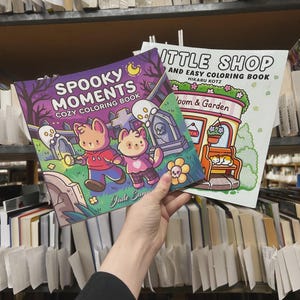 May include: Two coloring books are held up in front of a bookshelf. The cover of the first book reads "Spooky Moments Cozy Coloring Book" with illustrations of cartoon animals and ghosts. The second book is titled "Little Shop and Easy Coloring Book".