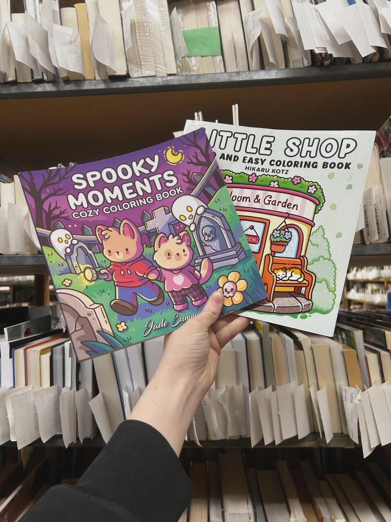 May include: Two coloring books are held up in front of a bookshelf. The cover of the first book reads "Spooky Moments Cozy Coloring Book" with illustrations of cartoon animals and ghosts. The second book is titled "Little Shop and Easy Coloring Book".
