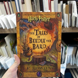 May include: The Tales of Beedle the Bard book by J.K. Rowling features a gold-toned cover with an illustration of a cauldron, a tree stump, and an axe. The title is printed in black text, and the Harry Potter logo appears at the top.
