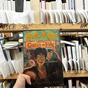 May include: A hardcover copy of "Harry Potter and the Goblet of Fire" by J.K. Rowling. The book's cover features an illustration of Harry Potter, with other characters and the title in gold lettering. The book is held in front of a shelf of other books.