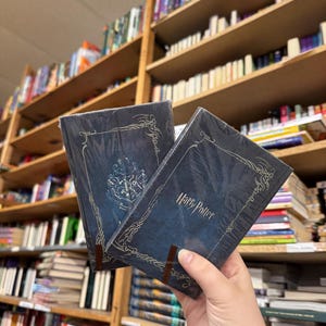 May include: Two copies of a "Harry Potter" book are held in front of a bookshelf filled with books. The books have a dark blue cover with gold decorative accents and the title "Harry Potter" in a script font.
