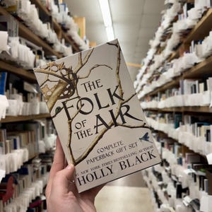 May include: A paperback gift set of "The Folk of the Air" by Holly Black. The book cover features a crown and branches design with the title text. The background shows bookshelves filled with books.