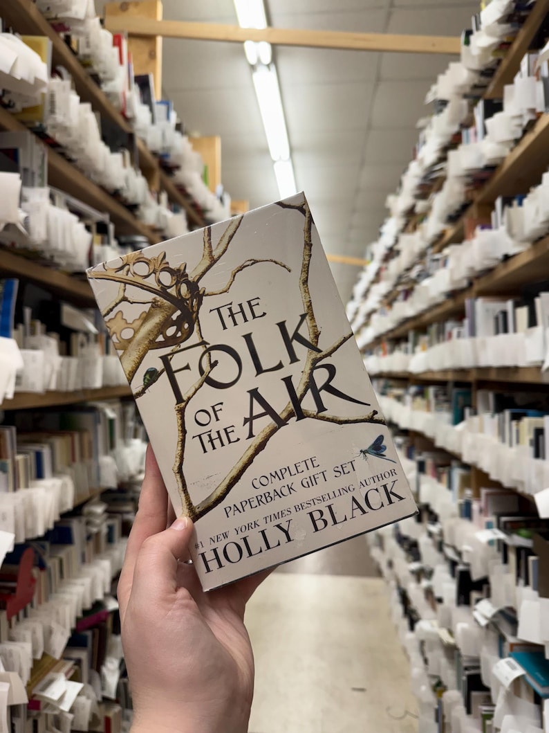 May include: A paperback gift set of "The Folk of the Air" by Holly Black. The book cover features a crown and branches design with the title text. The background shows bookshelves filled with books.