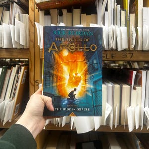 May include: A person holding a copy of "The Trials of Apollo: The Hidden Oracle" by Rick Riordan. The book cover features a vibrant illustration with a figure in silhouette against a fiery background. The book is surrounded by other books on a shelf.