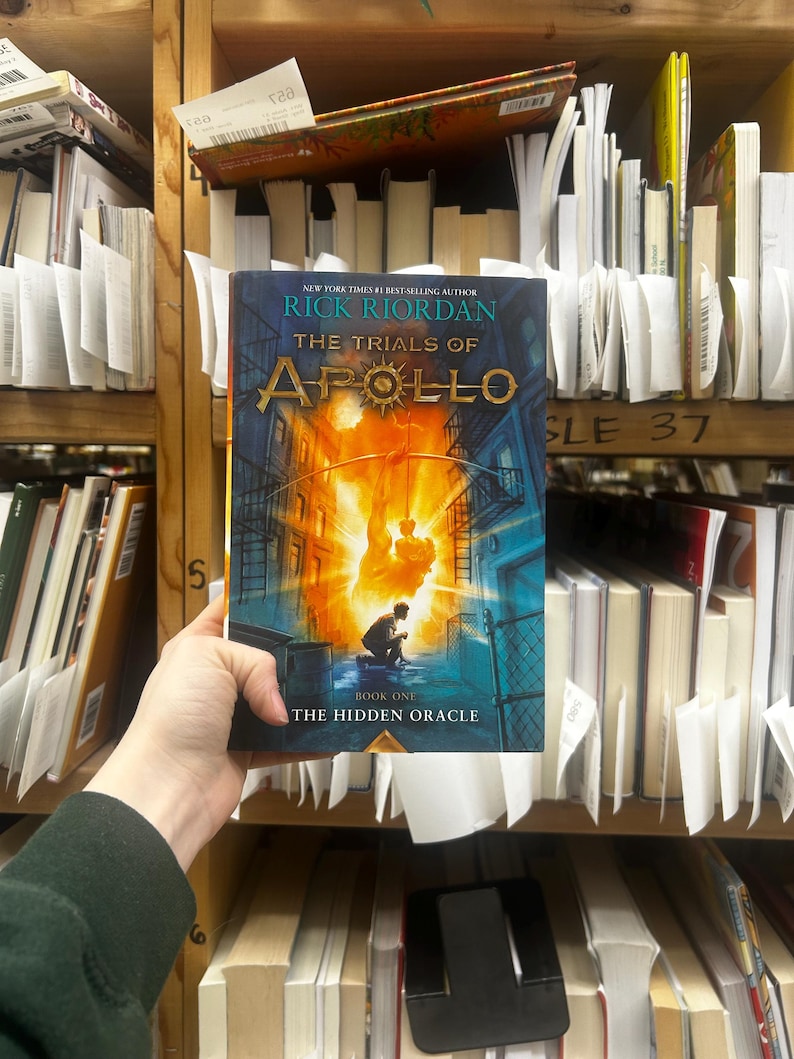 May include: A person holding a copy of "The Trials of Apollo: The Hidden Oracle" by Rick Riordan. The book cover features a vibrant illustration with a figure in silhouette against a fiery background. The book is surrounded by other books on a shelf.
