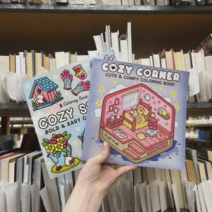 May include: Two coloring books titled "COZY CORNER" and "COZY S". The "COZY CORNER" book has a pink and blue cover with a bedroom scene. The "COZY S" book has a blue cover with a birdhouse and flowers. Both books are held in front of a bookshelf.