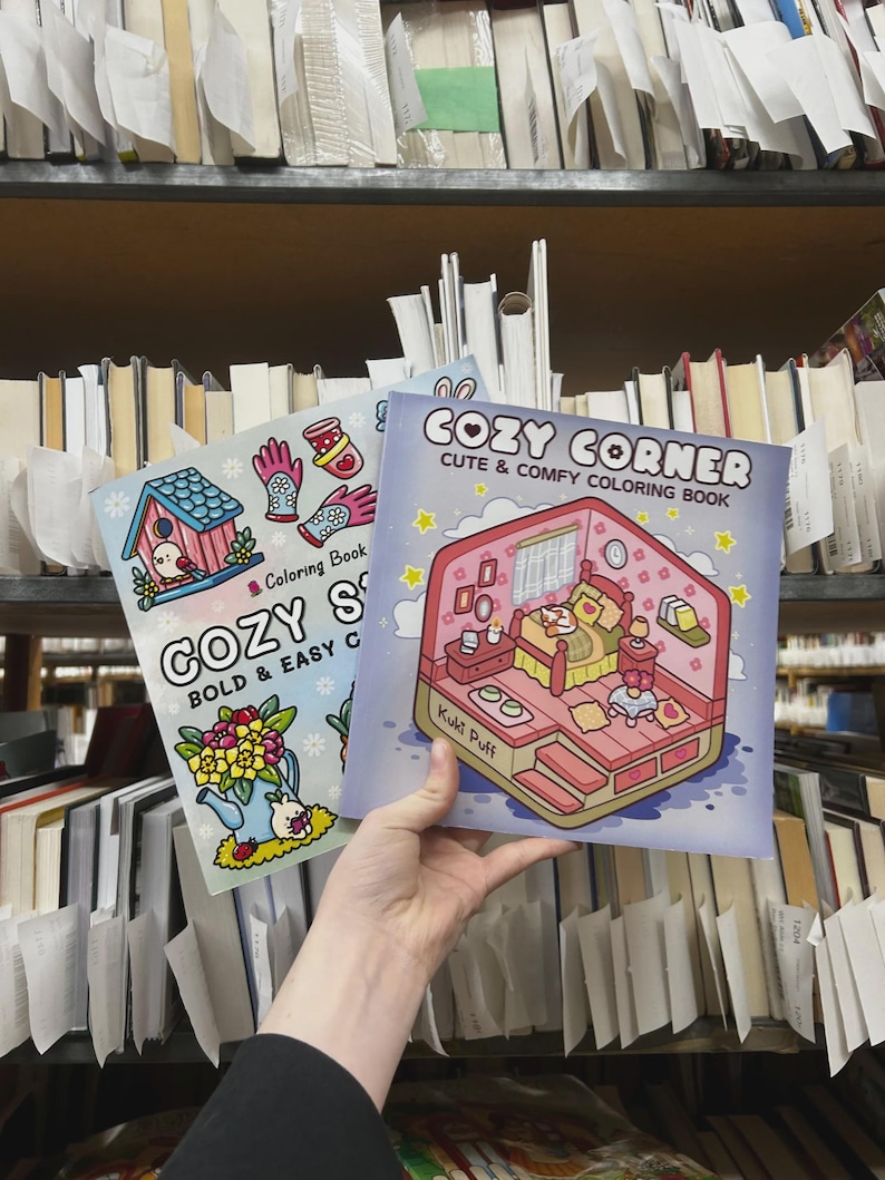 May include: Two coloring books titled "COZY CORNER" and "COZY S". The "COZY CORNER" book has a pink and blue cover with a bedroom scene. The "COZY S" book has a blue cover with a birdhouse and flowers. Both books are held in front of a bookshelf.