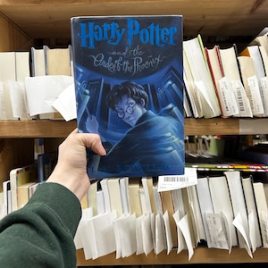 May include: A hardcover copy of "Harry Potter and the Order of the Phoenix" by J.K. Rowling. The book's cover features a blue and black illustration of Harry Potter. The book is held in front of a shelf filled with other books.