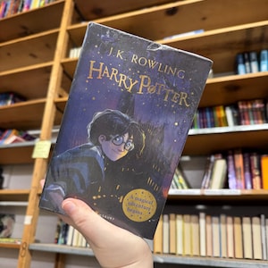 May include: A hardcover copy of "Harry Potter" by J.K. Rowling. The book cover features an illustration of Harry Potter with glasses, set against a dark blue night sky with gold lettering. The text "A magical adventure begins..." is also visible.