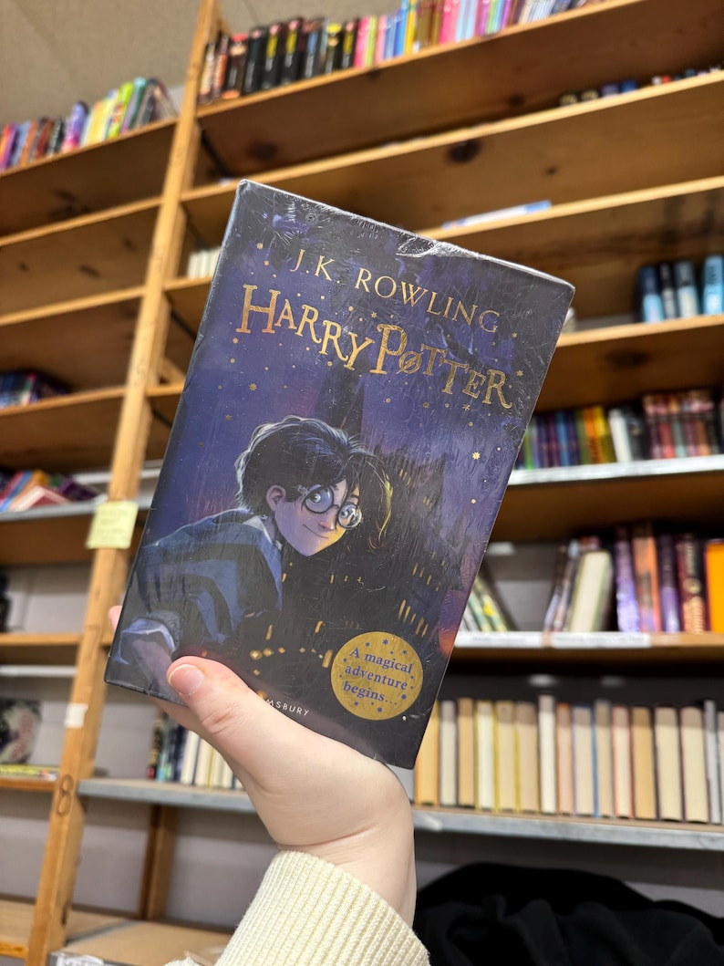 May include: A hardcover copy of "Harry Potter" by J.K. Rowling. The book cover features an illustration of Harry Potter with glasses, set against a dark blue night sky with gold lettering. The text "A magical adventure begins..." is also visible.